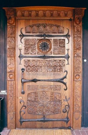 A wooden door intricately carved with ornate patterns, figures, and symbols, featuring decorative metal accents and a circular detail at the center. The door has a rustic and artistic appearance.