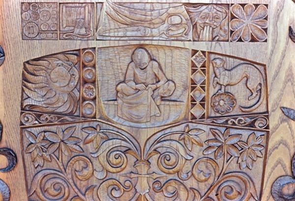 Intricate wooden carving depicting a central figure sitting cross-legged, surrounded by geometric patterns, floral motifs, a horse, a sun, and abstract designs.