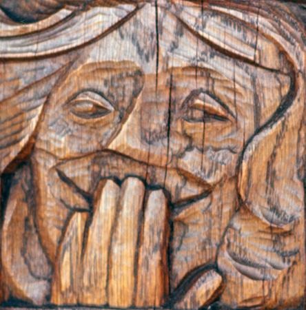A wooden carving depicts a face with closed eyes and long hair, holding several fingers to its mouth in a gesture that may suggest laughter or surprise. The carving shows intricate detail and texture in the wood.