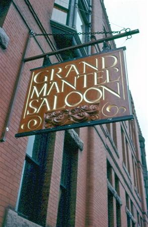A hanging sign reading "Grand Mantel Saloon" in gold letters is attached to a brick building, with windows and more brickwork visible in the background.