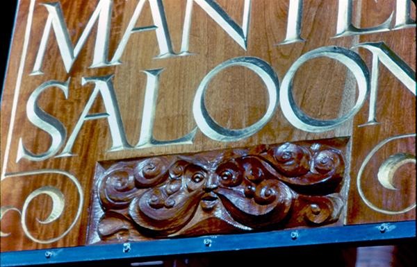 A close-up of a wooden sign reading "SALOON" with ornate carved decorations below the letters and part of another word visible above. The lettering is painted in a light color on varnished wood.