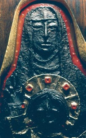 A textured, dark-toned relief sculpture depicting a solemn-faced figure draped in a veil, holding a smaller circular figure with raised hand; red and gold accents highlight the artwork.