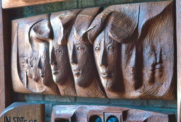 A wooden relief carving depicts five abstract human faces in profile, closely aligned side by side, with smooth, stylized features and textured hair against a brick background.
