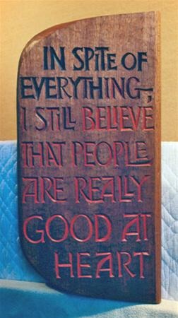 A wooden plaque with engraved text reads: "IN SPITE OF EVERYTHING, I STILL BELIEVE THAT PEOPLE ARE REALLY GOOD AT HEART." The words "GOOD AT HEART" are highlighted in red. The plaque rests against a quilted surface.