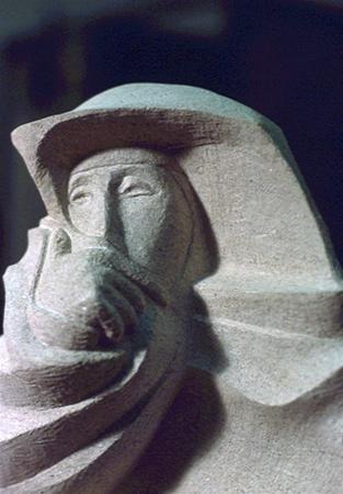 A close-up of a stone sculpture depicting a person wearing a hood or head covering, with a solemn expression and stylized, abstract facial features.