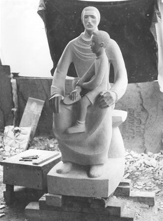 A stone sculpture depicting a seated adult figure with a child on their lap, set in a workshop with tools and debris on the ground. The background is partially covered with fabric.