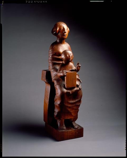 A wooden sculpture of a stylized woman standing with a child on her lap. Both figures have abstract, rounded features and the child is holding a rectangular block. They stand on a square base against a plain background.