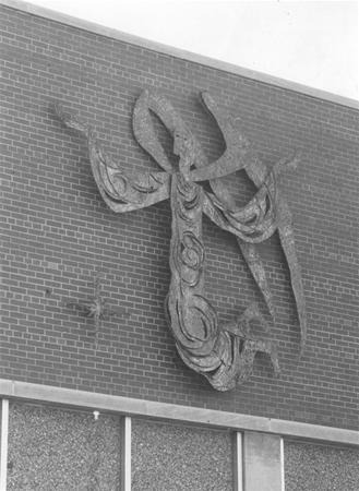 A metal sculpture of an angel with outstretched arms is mounted on a brick exterior wall above windows. The angel has swirling patterns in its robe and a halo around its head.