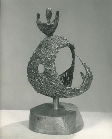 A textured abstract bronze sculpture features a hollow, curved form with rough surfaces, mounted on a cylindrical base. At its top, a smaller human-like figure with raised arms emerges, adding contrast to the flowing shape.