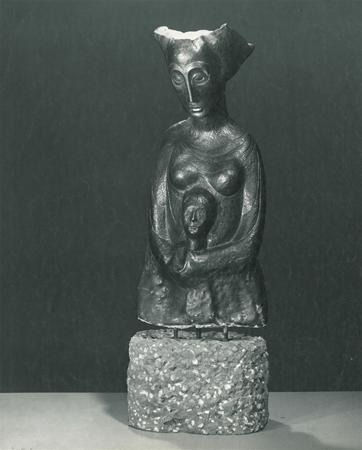 A black and white photo of a stylized sculpture depicting a seated female figure holding a smaller human figure on her lap, mounted on a textured stone base.