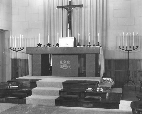 Black and white photo of a church altar with a crucifix, candles on tall stands, and a fish symbol on the altar front. Steps with kneeling pads lead up to the altar, and curtains hang behind it.
