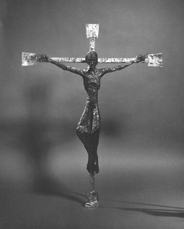 Black and white photo of a modern, abstract metal sculpture of Jesus on the cross, with elongated arms and a textured, rough surface, set against a plain background.