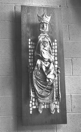 Black and white photo of a relief sculpture depicting a crowned figure seated on a throne, mounted on a rectangular wooden panel, with a brick wall in the background.