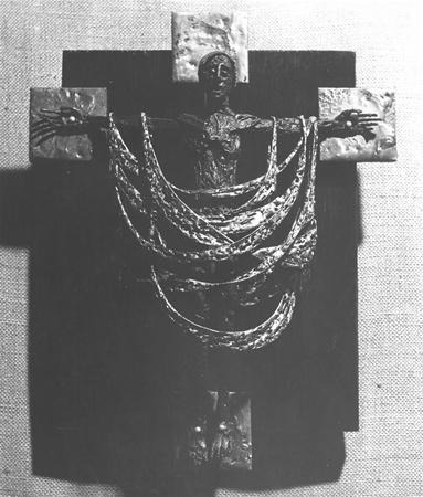 A sculpted crucifix features a figure with outstretched arms, draped in textured, leaf-like patterns, mounted on a dark cross with raised square ends, against a neutral fabric background.