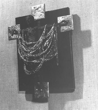 A sculpted metallic crucifix features a figure of Christ with draped, textured cloth and outstretched arms, mounted on a dark wooden cross against a light fabric background.