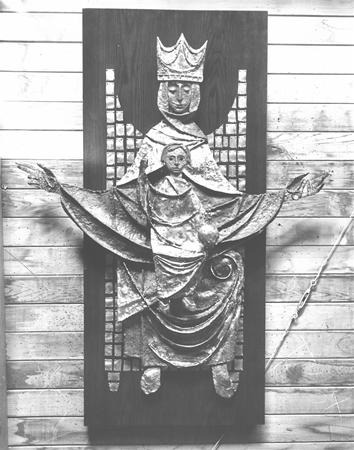 A metal sculpture of a crowned figure holding a child with outstretched arms, mounted on a wooden panel with a grid-like background, displayed against a wooden wall.