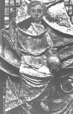 A metallic relief sculpture depicts a stylized human figure holding a spherical object, with draped, textured clothing and abstract, angular shapes in the background. The image is in black and white.