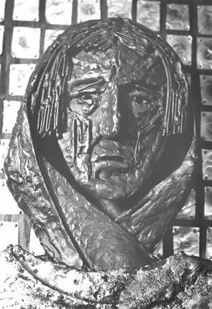 A grayscale photo of a textured metal sculpture depicting a sorrowful face wrapped in a headscarf, set against a grid-patterned background. The expression appears solemn and somber.