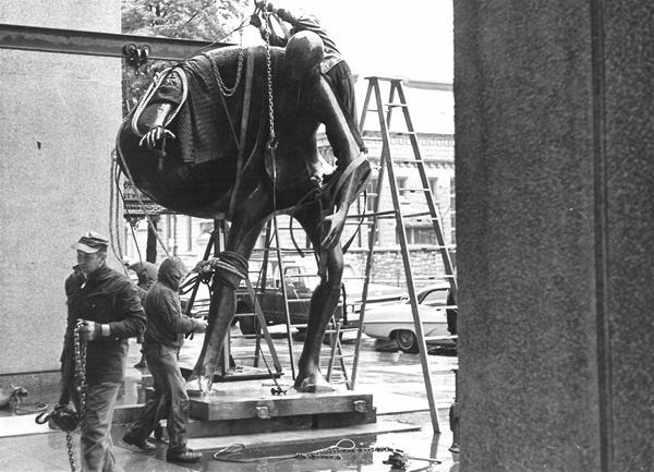 Black and white photo of workers installing a large, chained sculpture of a horse and rider outside, with ladders and parked cars visible in the background.