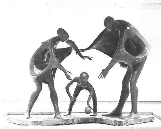 Three abstract, elongated bronze figures are arranged in a dynamic pose, with two taller figures leaning over a smaller figure in the center who is bending down to touch a ball. The figures have smooth, stylized forms.