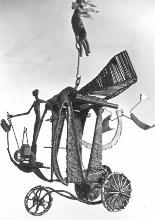 A black and white photo of a whimsical metal sculpture featuring a winged figure on a wheeled cart, with abstract shapes and a suspended figure holding objects above.