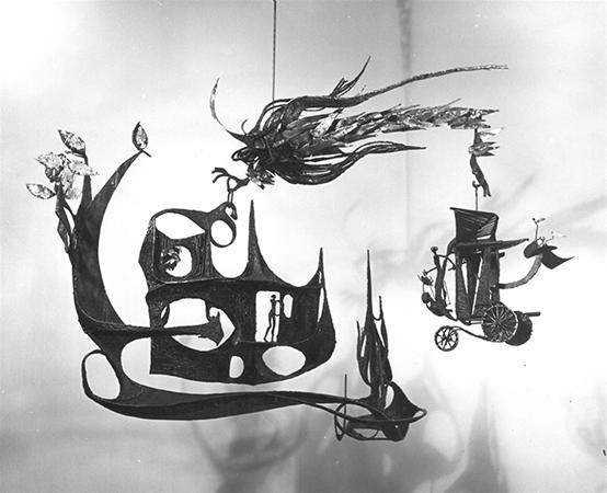 A black-and-white photo of an abstract, whimsical mobile sculpture with organic shapes, silhouettes of people, a tree, a coach with wheels, and fantastical creatures hanging in midair.