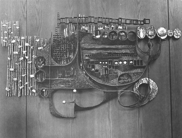 Black and white photo of an abstract metal wall sculpture featuring geometric and circular shapes, grids, and textured surfaces, mounted on wooden panels. The artwork has an industrial, intricate design.