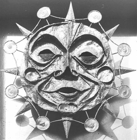 A metallic sun mask with a smiling human face, surrounded by rays and smaller circular shapes connected around its perimeter. The mask has exaggerated eyes, nose, and lips, and is in black and white.