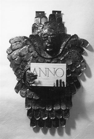 Black and white photo of a metallic angel sculpture with detailed wings, holding a rectangular sign that reads "ANNO," with three small silhouettes at the bottom of the sign.