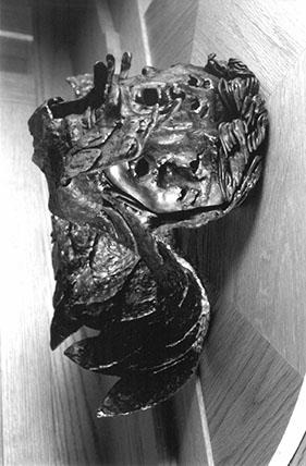 A black-and-white photo of an abstract bronze sculpture mounted on a wooden wall, featuring textured surfaces, organic shapes, and what appears to be a partial human face and hand emerging from the form.