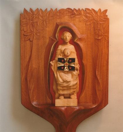 A wooden relief carving depicts two robed figures, one standing behind the other, with a decorative shield adorned with red and black elements. Leaf patterns are carved above them on the wooden background.