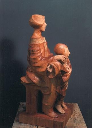 Wooden sculpture of a seated woman with a child standing between her legs, both wearing draped clothing, set against a dark background on a wooden surface.