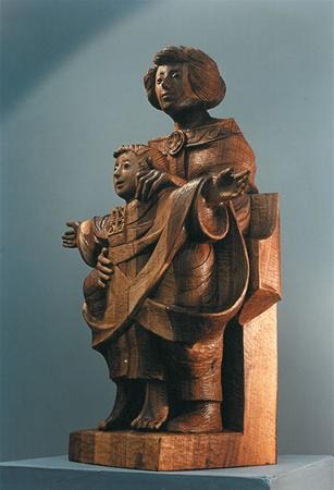 Wooden sculpture of two figures, an adult and a child, standing closely together. The adult holds the child with one arm and drapes a robe around them; both figures have serene expressions and detailed clothing.