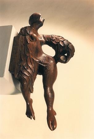 A wooden sculpture of a human figure with bird-like features, including carved wings and webbed feet, mounted on a wall and looking upward. The figure has an abstract, flowing design.