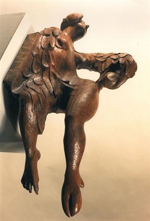 A wooden sculpture of a winged, human-like figure is attached to a wall, sitting with one leg extended downward and the other bent, holding an object in its right hand. The figure appears to be in motion.