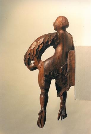 A wooden sculpture of a human figure with bird-like wings and hoofed feet is attached to the side of a rectangular block against a plain background. The figure appears to be looking upward and to the side.