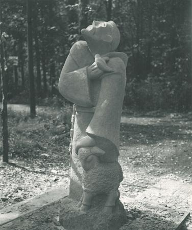 Stone sculpture of a standing figure in a robe holding a lamb over its shoulders, looking upward. The background is a wooded outdoor area with sunlight filtering through the trees.