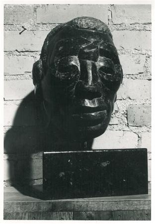 Black stone sculpture of a stylized human head with abstract features, displayed on a rectangular base in front of a brick wall.