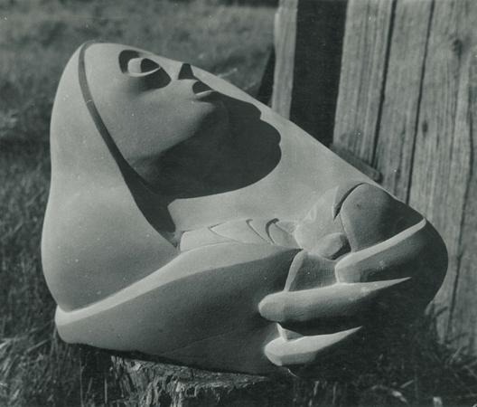 Black and white photo of a smooth, abstract stone sculpture depicting a partially reclining human figure with an upward-facing head and stylized hands, set outdoors against wood and grass.