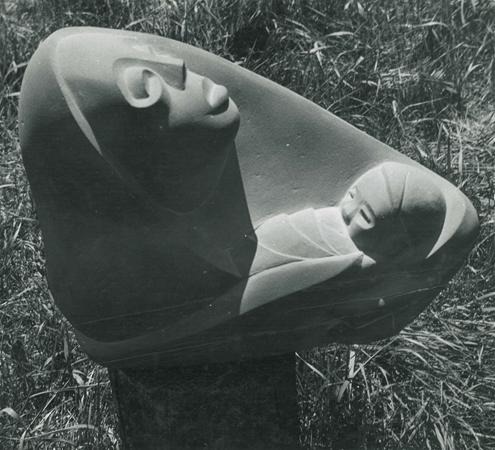 A stone sculpture depicting a stylized adult face looking upward and a swaddled baby, both carved from the same block, is displayed outdoors on grass.