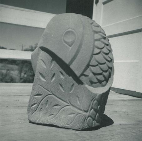 A carved stone sculpture resembling a stylized fish with scales and leaf patterns sits on a wooden floor near an open door and a railing in the background.