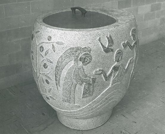 A large stone urn with a metal lid, decorated with carvings of human figures, birds, and plants against a brick wall background.