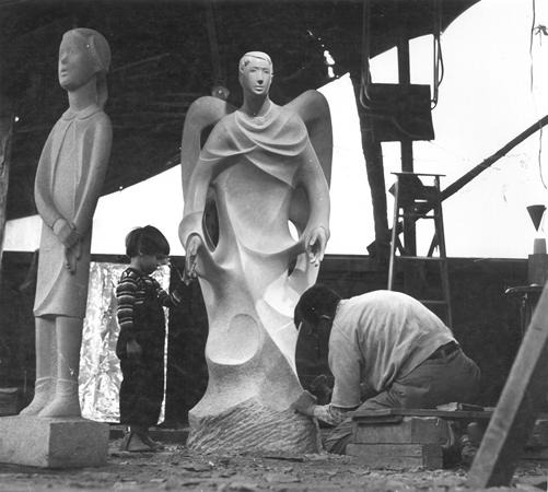 A sculptor kneels while working on a large abstract stone sculpture. A child stands nearby looking at another finished sculpture of a standing figure. The scene is set in a workshop with tools and ladders visible.