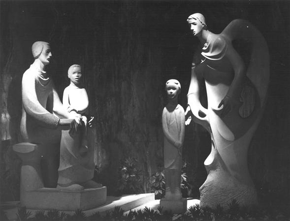 Black-and-white photo of four abstract, smooth stone statues—two adults and two children—positioned in a dimly lit, natural setting with plants at their base, appearing to interact with one another.