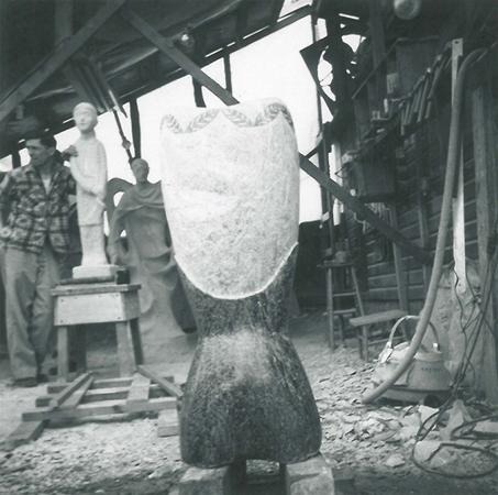 A black-and-white photo shows an abstract stone sculpture in the foreground, with two men and two figurative sculptures in the background, all within a workshop filled with tools and equipment.