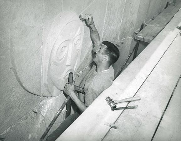 A man uses a chisel and mallet to carve a face with large eyes and a round mouth into a stone wall. Several tools are scattered on a plank beside him.