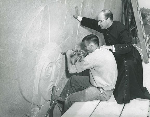 A man carves a large face into stone while another man in clerical clothing observes, resting one hand on the wall beside the carving. They are working on scaffolding.