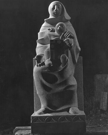 A stone sculpture of a seated, robed woman holding a child on her lap. The figures have simplified, abstract features and the woman holds a rectangular object in one hand. The background is dark.