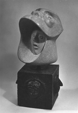 Black and white photo of an abstract sculpture featuring a stylized human face with a large, curved, helmet-like form above it, mounted on a square base with embossed designs.
