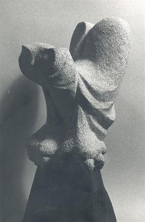 A textured stone sculpture depicting an abstract, draped figure with flowing, wing-like shapes, positioned against a plain background and illuminated to highlight its intricate surface.